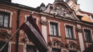 Latvijas valsts karogs plīvo pie vēsturiskas ēkas Rīgas centrā, simbolizējot nacionālo piederību.