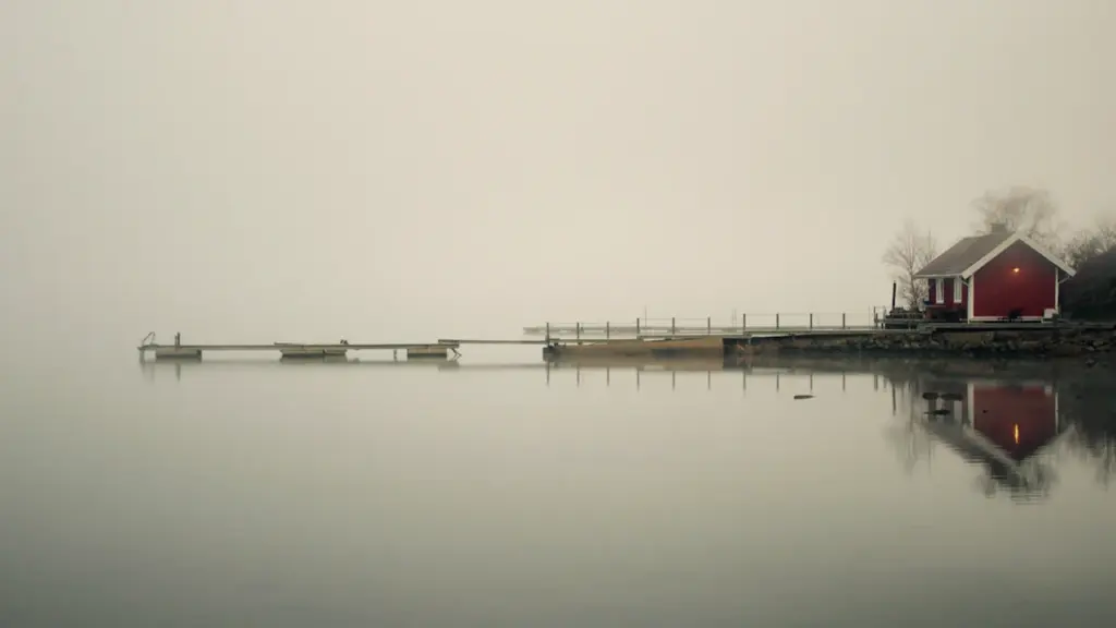 Mierīga sarkana koka mājiņa pie piestātnes miglainā rīta ainavā pie ezera krasta.