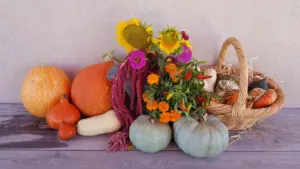 Krāsains rudens ražas grozs ar ķirbjiem, saulespuķēm un rudens ziediem uz galda.