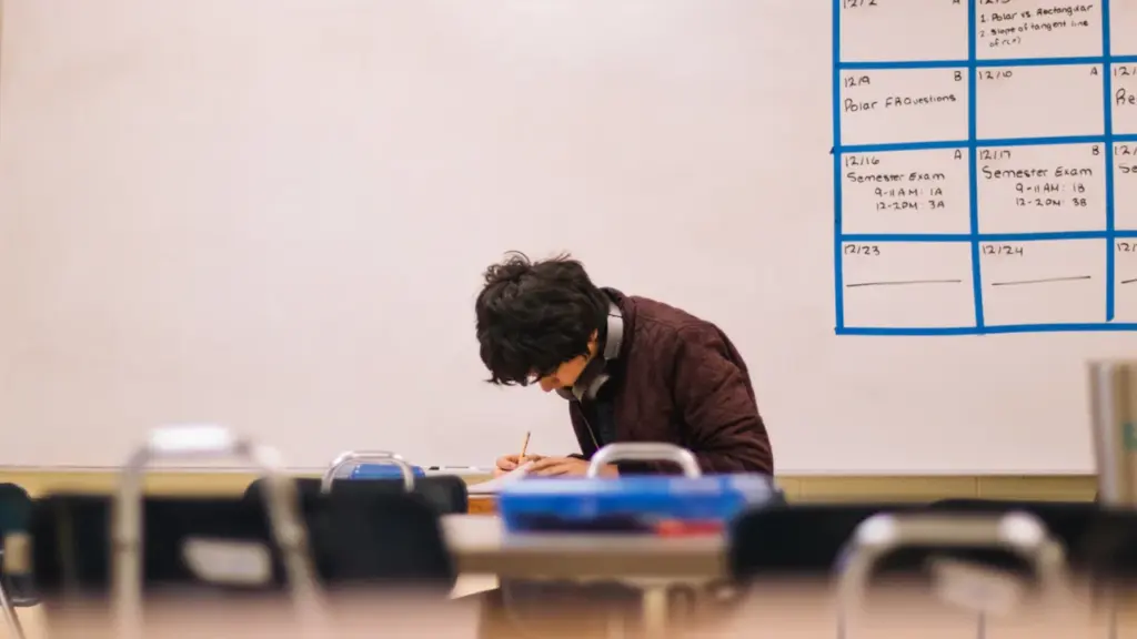 Students koncentrēti strādā pie rakstāmgalda klases telpā ar tāfeli un grafiku fonā.